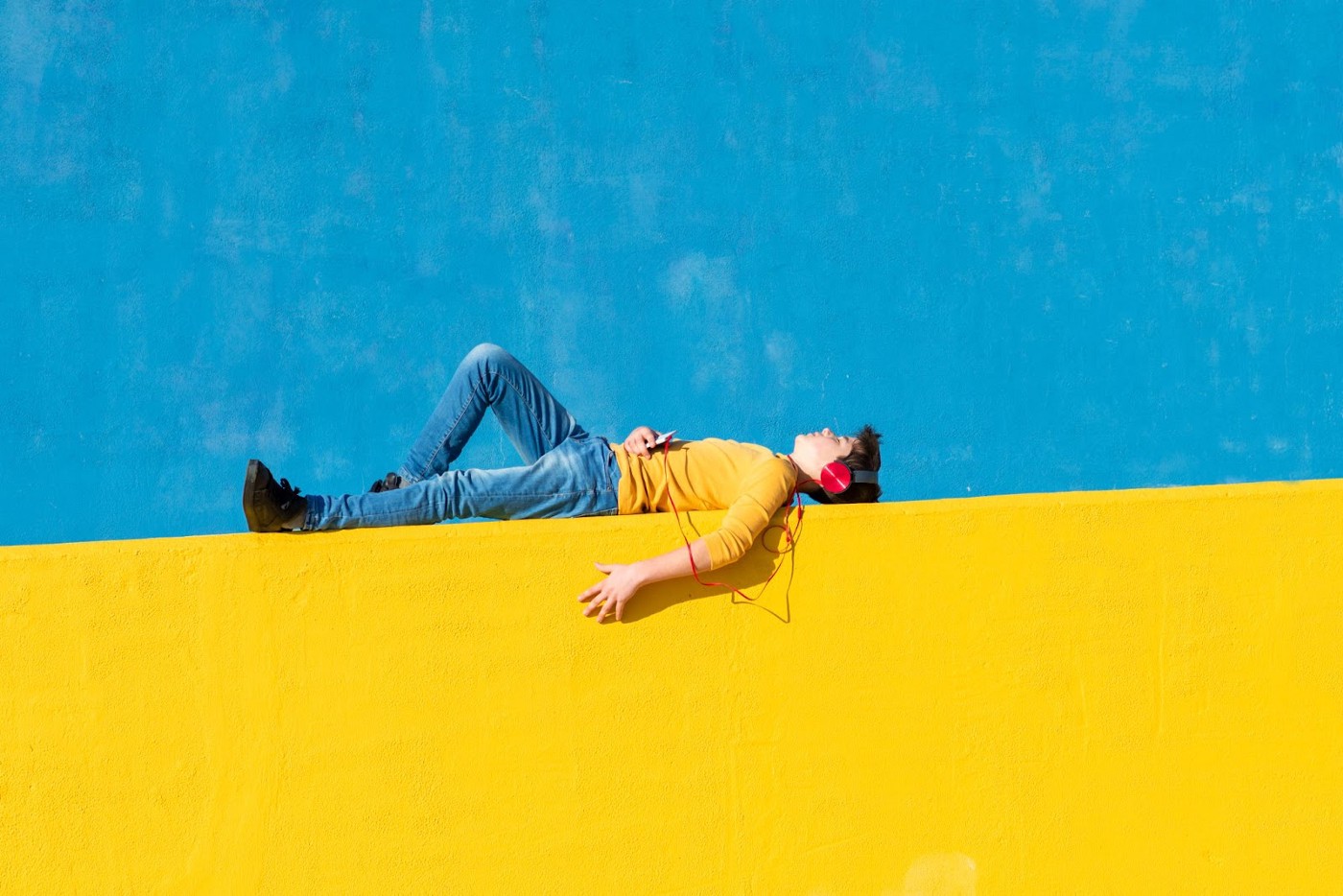 Creativity beats aggression: Boy with headphones lying on a yellow wall with blue background