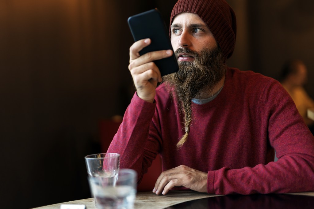 Adult man with a braided beard using the Earkick app on his smartphone to do a self-check-in via voice recording. He has a glass of water in front of him and is looking reflective and vulnerable. Having a way to express his emotions helps him protect his mental readiness and disconnect from worries efficiently.