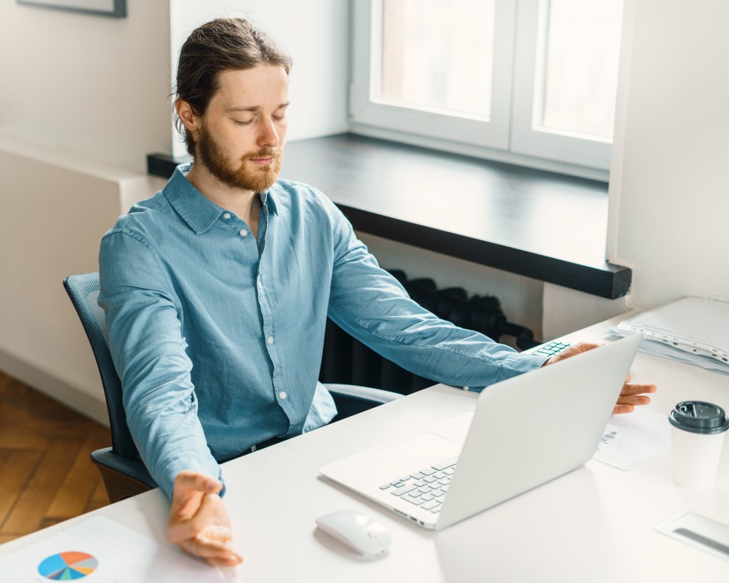 Relief stress at work. Calm male office worker with closed eyes doing breathing exercise, keeping hands in mudra while sitting at desk with laptop in office. 