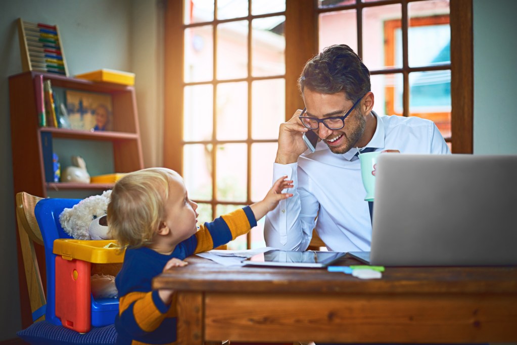 Relaxed businessman working from home, taking a call while his toddler enjoys his company. The father is drinking his tea and smiling at his little boy. Being close to his family not only saving him time but also keeping his mental health in check.