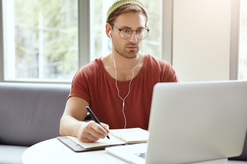 Mateo in front of his laptop, focusing on his goals. To deal with his style of perfectionism he's prioritizing his tasks of the day in his pen-and-paper agenda, keeping distractions away with headphones in ears.