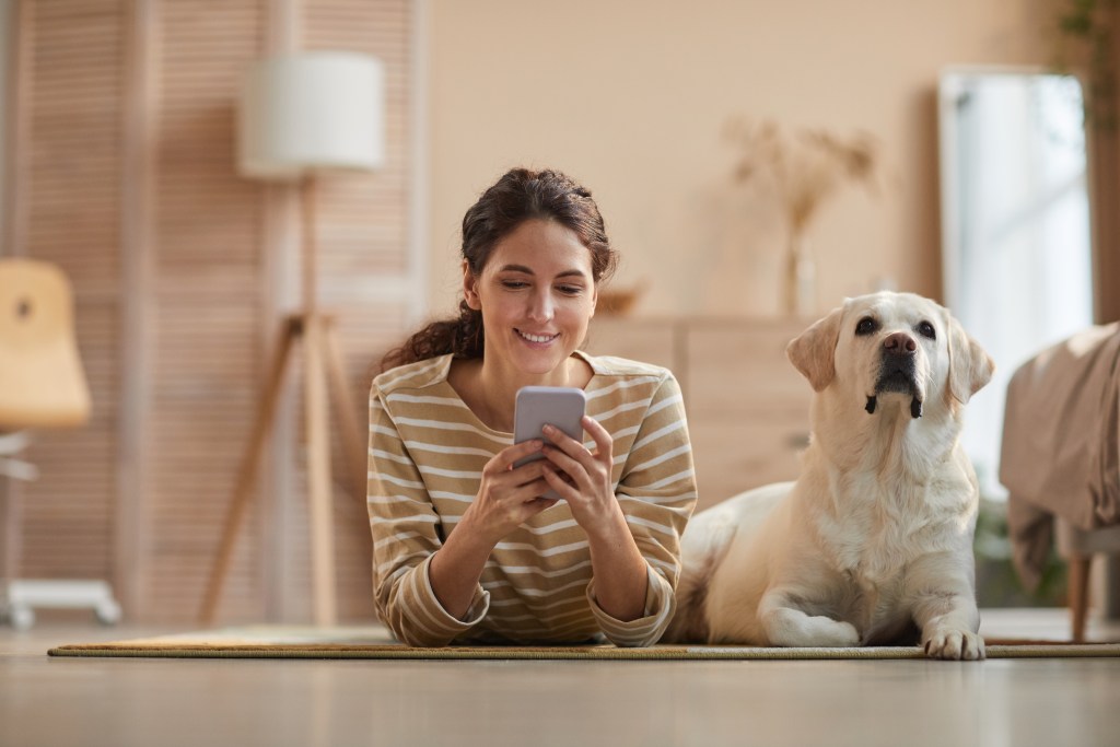 Front view portrait of smiling young woman lying on floor with her dog and using the Earkick self-care companion app to do a self-check-in and check her stats. Since she's decided to have a dog, her mental readiness and mental health have improved visibly.