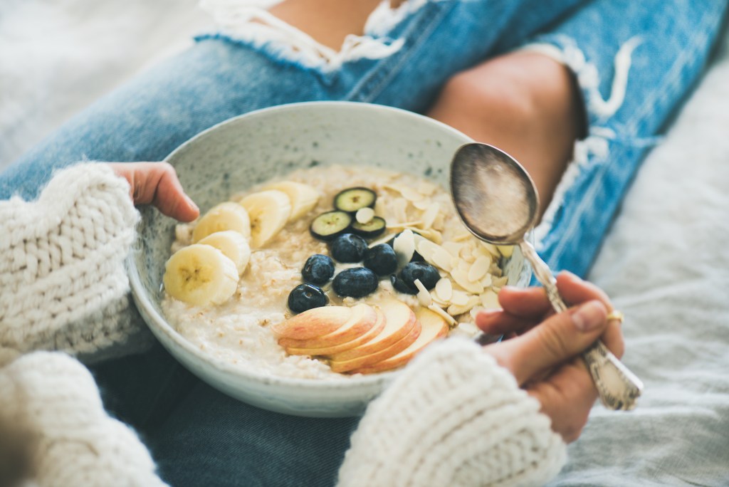 Selfcare routines and healthy habits: Woman in jeans and wool pullover taking time for a healthy snack at work. She holds a bowl of oats, fruits, joghurt and almonds on her lap and a spoon in her hand.