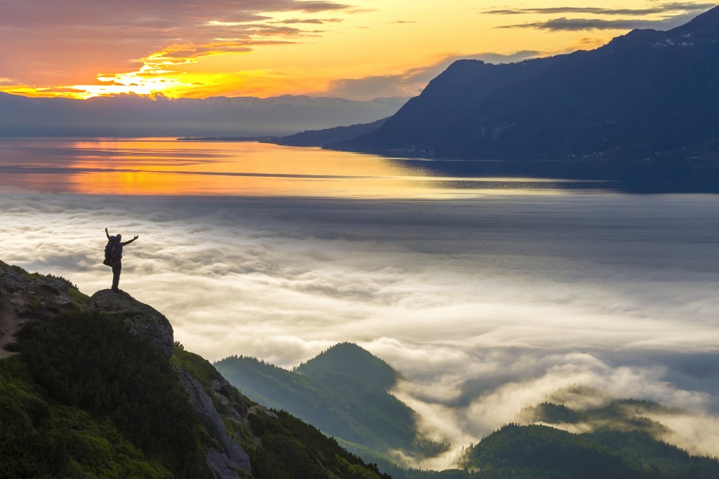 Small silhouette of a person with backpack on rocky mountain slope with raised hands over valley covered with white puffy clouds, watching the sun rise.