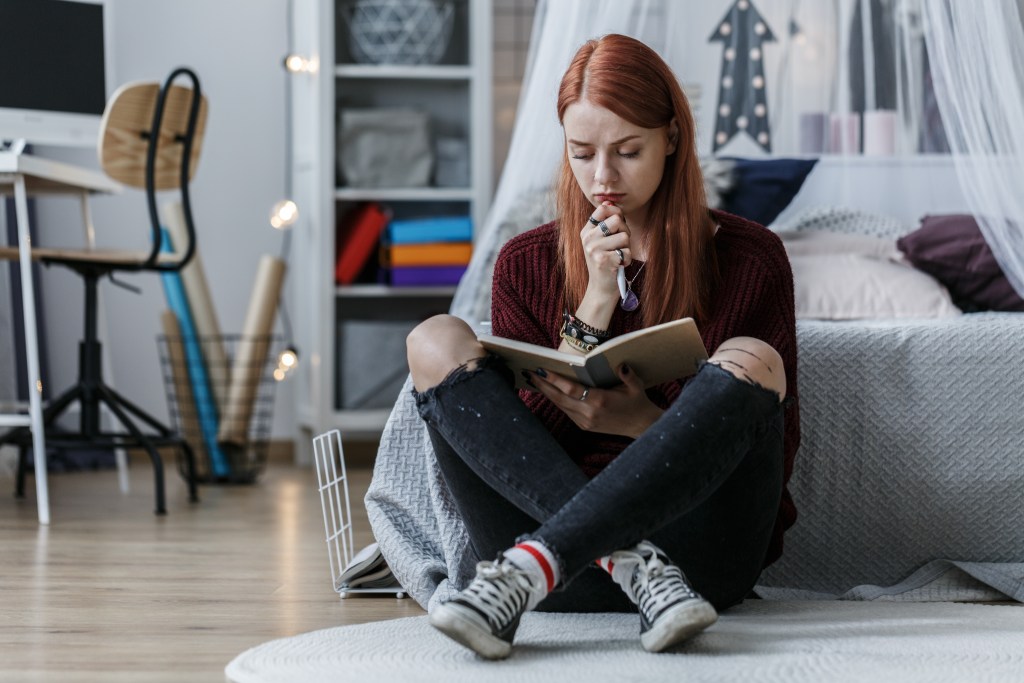 Young red-haired woman sitting next to her bed and journaling. She's reflecting about her day's events, capturing