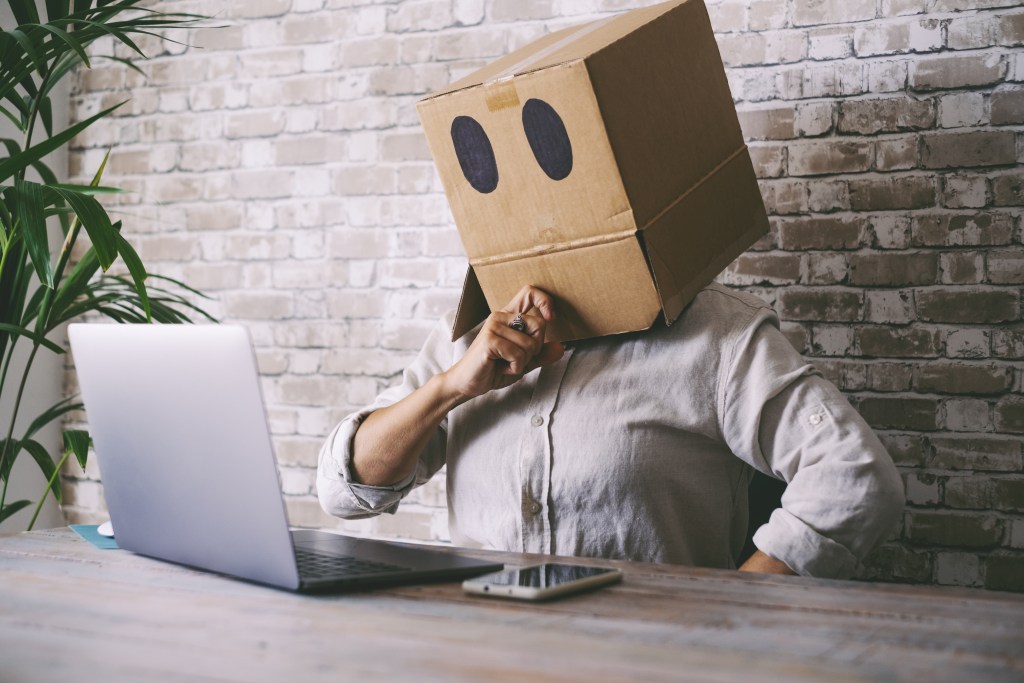 Woman hiding her face in a cardboard box while sitting in front of her computer. It's Finance professional Trisha who hides her struggles from managers & colleagues: A common case of the health-privacy paradox.