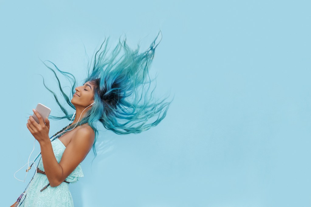 Exercise made easy: Cheerful young woman with flying blue hair listening to music on her phone and dancing while going for a short stroll outside.