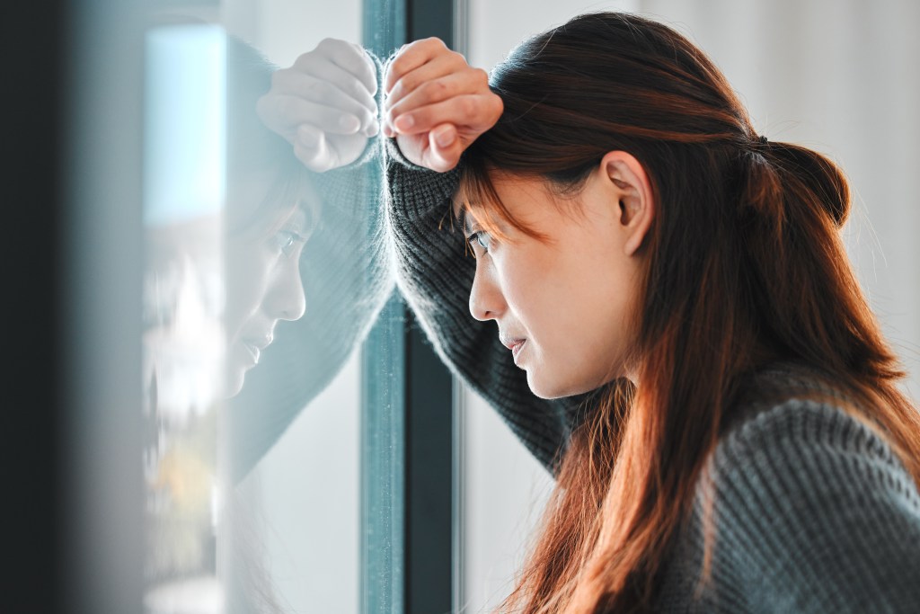 Young female professional ruminating about a work-related problem, feeding the cycle of anxiety. She's leaning her head against her arm, looking out the window without really taking in what is going on in front of the window.