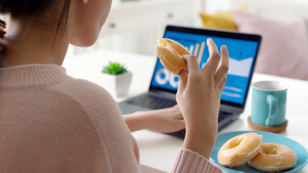 Young female professional coping with stress and anxiety by snacking doughnuts while going through her presentation on the computer in front of her.