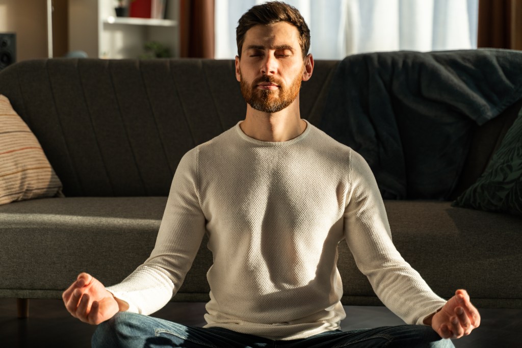 Mindfulness in the home office: Male business professional in lotus pose and with closed eyes, meditating to disconnect from anxious thoughts.
