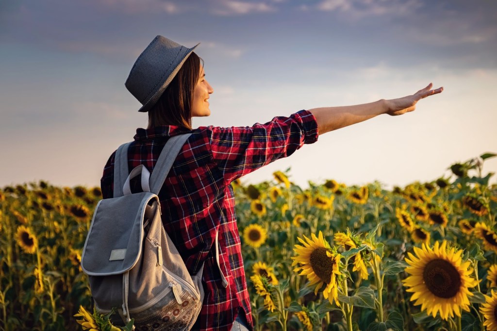 Smiling young woman in a sunflower field, extending her right arm towards the sun and capturing the moment with all her senses, feeling 100% alive.