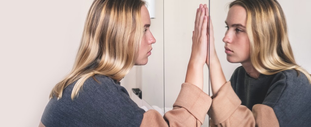 Female blond student looking at herself in a mirror, with her hand on the mirror. She's feeling cynical, doubting herself, others, her work and her life.
