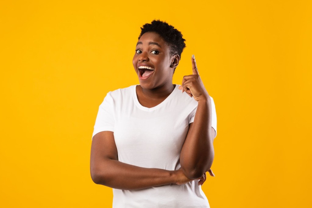 Eureka Moment: Cheerful Black Woman Pointing Finger Up Having Great Idea Posing Smiling To Camera Standing On Yellow Studio Background. She had a great idea after sleeping well and letting her brain process information thoroughly.