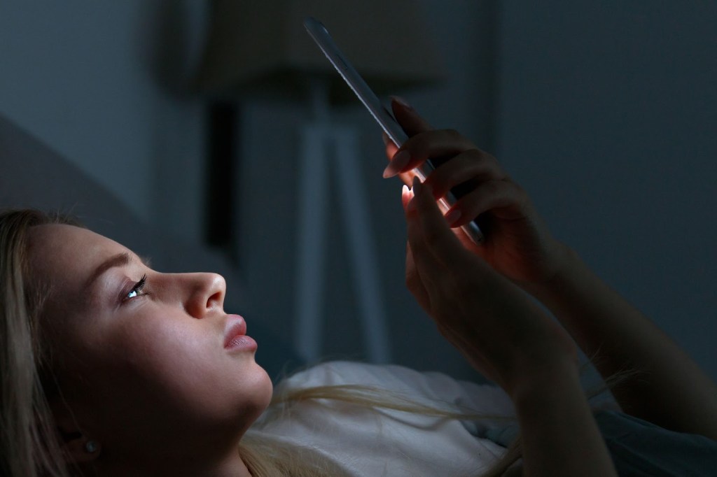 Young woman chatting and surfing on internet with smartphone late at night, lying on bed under the blanket, losing out on sleep due to social network addiction and nomophobia.  
Social network addiction, nomophobia, insomnia, sleep disorder concept. Bored woman chatting and surfing on internet with smartphone late at night, lying on bed under the blanket.