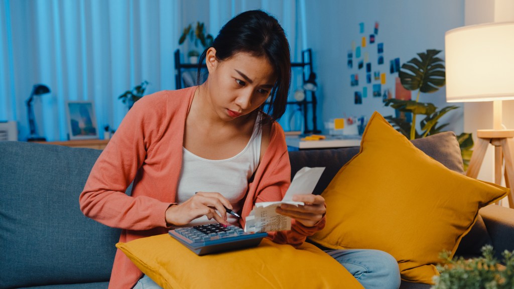 Budgeting and bills can be anxiety-inducing: worried Asian-American teenage girl sitting on a sofa and calculating her expenses.