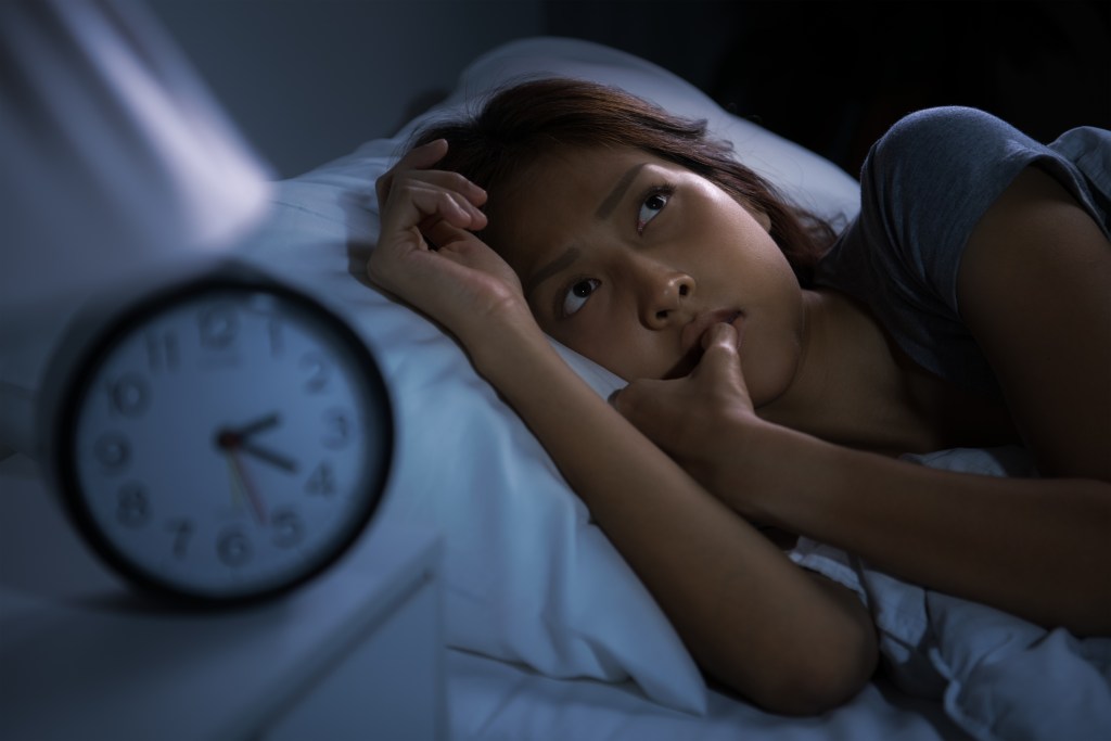 Worried young Latina woman in bed, struggling with racing thoughts at night. Next to her is an alarm clock showing 2:20 am