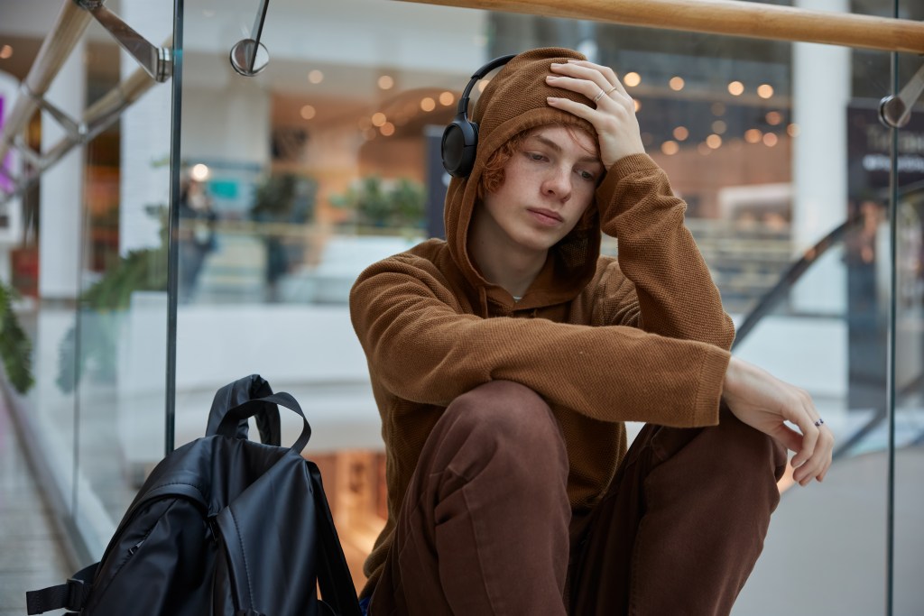 What does burnout feel like? Male red-haired student sitting nearby moving staircase at shopping mall. He looks depressed, can't muster up the energy to join peers. He has his headphones up and is listening to music while feeling negative emotions.