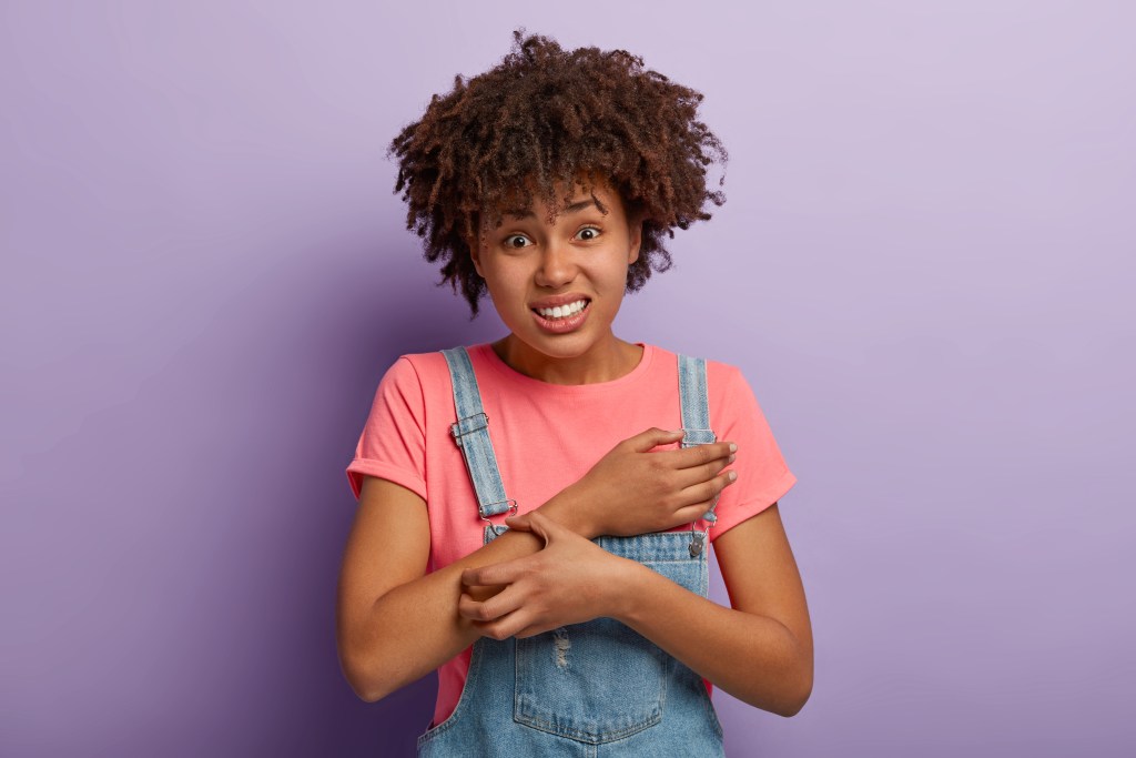 Displeased curly African American woman scratches her annoying stress rash on her right arm. It was caused by too much pressure during exam period.
