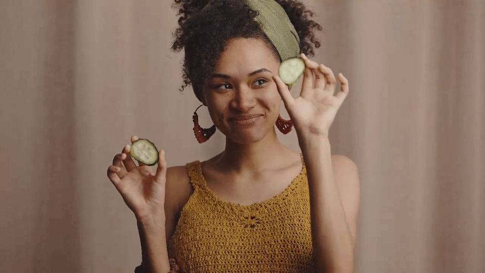 Young smiling African-American woman putting slices of cucumber on eyes on her self care Sunday