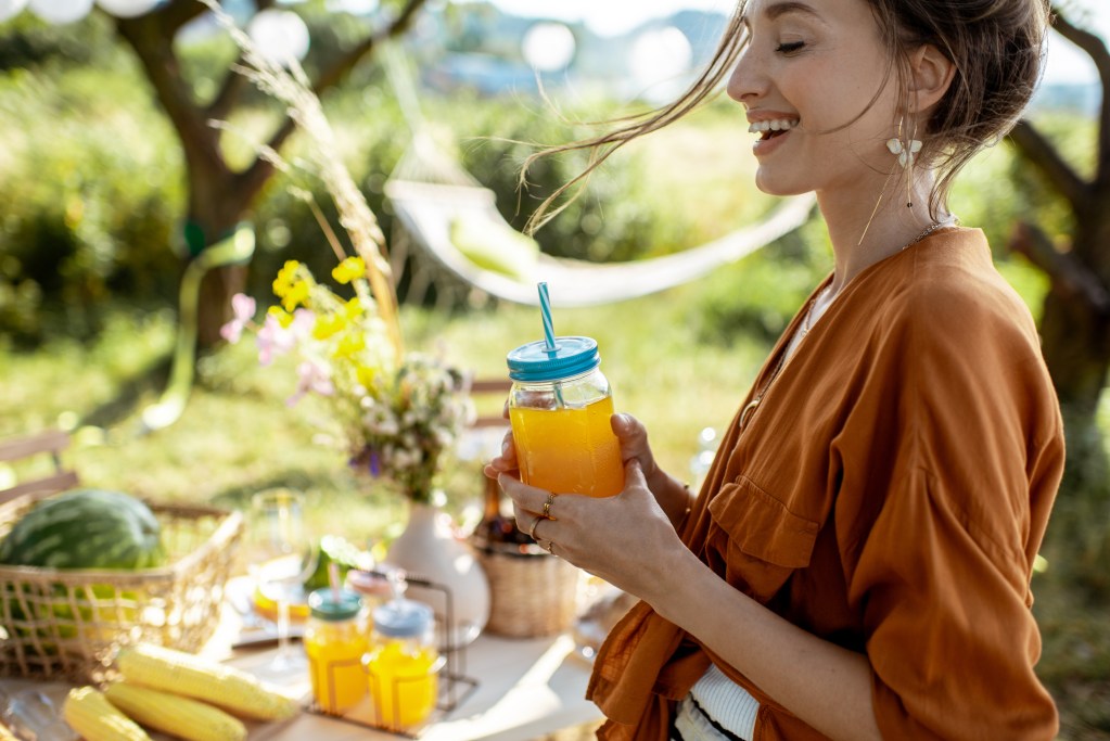 Caucasian woman laughing and holding a jar with juice and straw in her hands, standing at a beautifully decorated brunch table in the garden