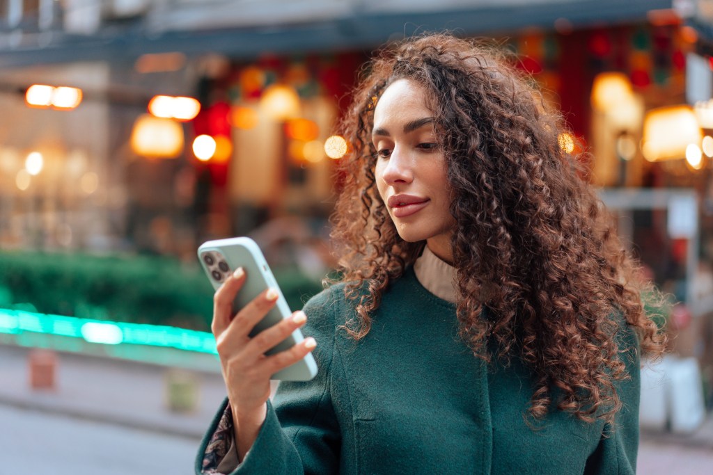 Young woman, student with curly hair holding her smartphone and testing personal AI chatbot for mental health to manage her anxiety after a previous negative experience in traditional therapy

