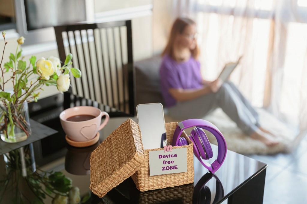 Screentime under control: Young woman sitting in the background, reading a book in her device free zone. In front there is a box that contains her smartphone, headphones and all digital devices. Concept of mental and digital detox.
