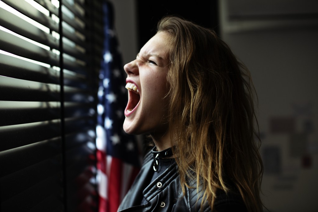 Election anxiety: Young woman in front of a window with the blinds drawn, next to an American flag, screaming with frustration over uncertainty and political stress