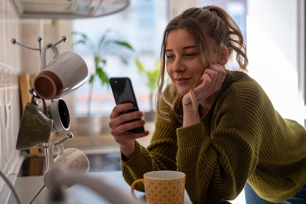 Young woman using one of the AI companions on her smartphone while standing in the kitchen.