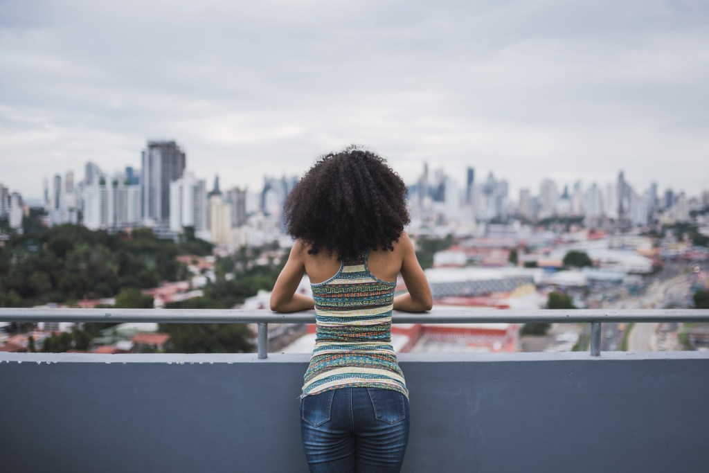 Back view of  young woman with long curly hair standing on balcony looking at the new city she moved to. She feels the separation anxiety in adults first hand.