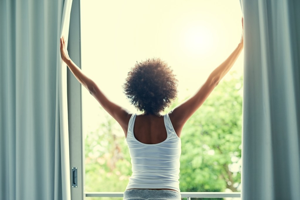 Rising like a sleep goddess: Portrait of assertive and rested woman opening the curtains in her bedroom at home after a good night's sleep