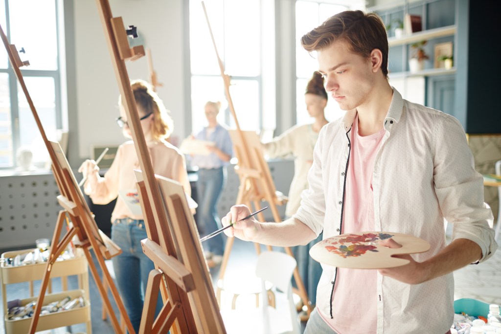 Prone to creative burnout: serious guy concentrating on his painting on easel during lesson in school of arts