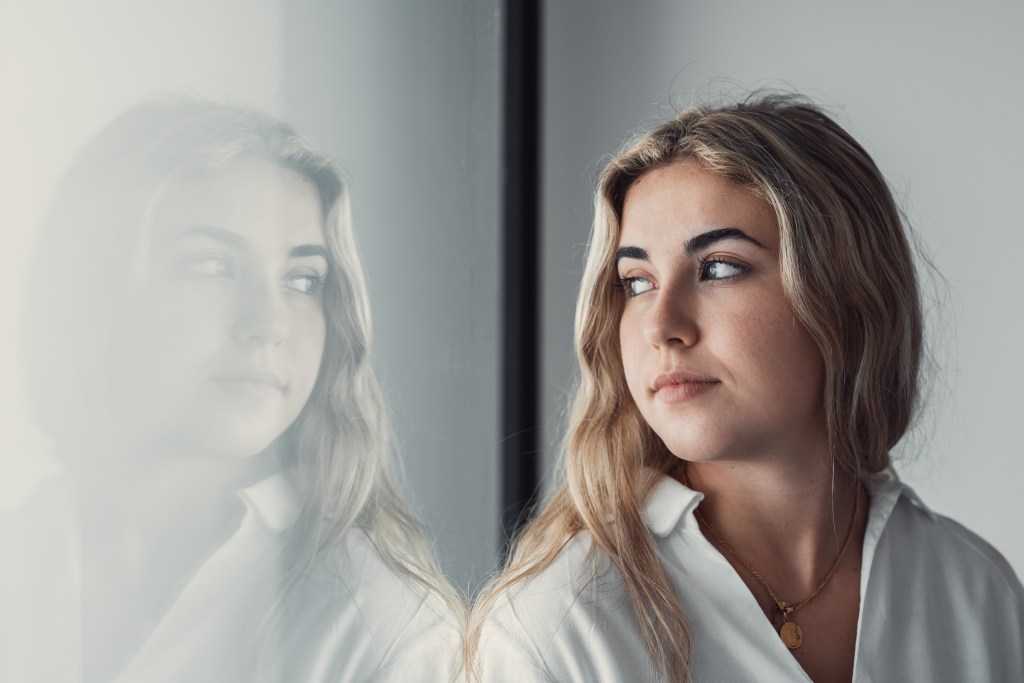 "I don't feel like myself anymore": Woman looking over her shoulder at her reflection in the window. She's doubting herself, looking pensative almost sad.