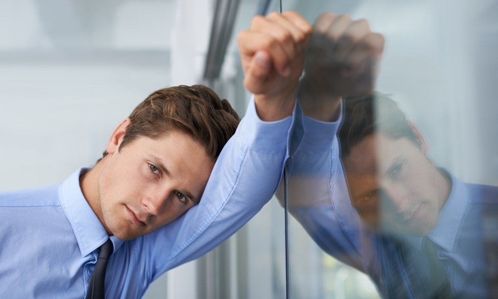 Performance anxiety at work: Tired young businessman leaning against a glass wall