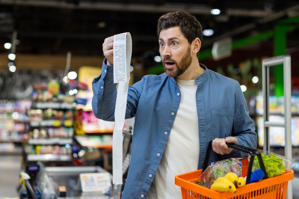 Tariff anxiety: Shocked man with rounded eyes and open mouth looking at unusually high prices while holding orange food basket.