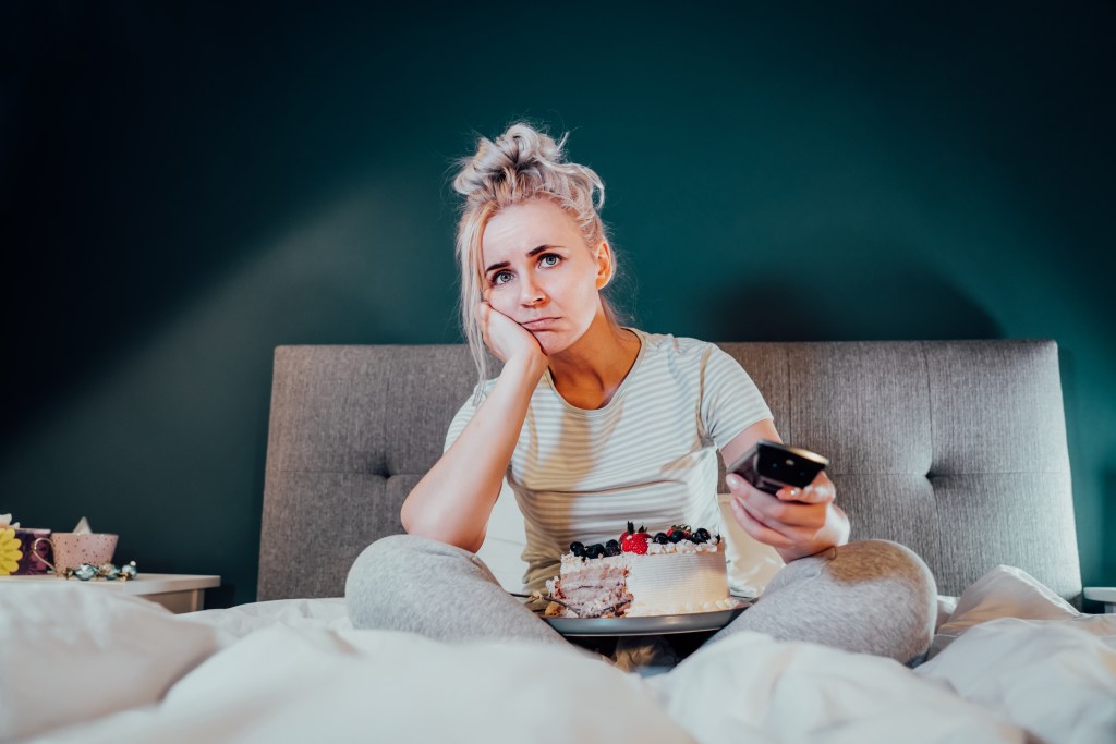Bored, sad woman in home clothes eating cake and using the remote control, bed rotting