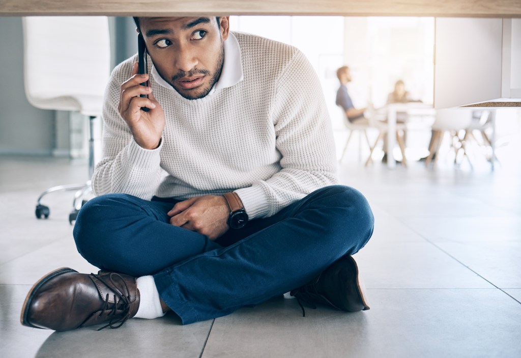 Man making frantic phone call, hiding under desk, not knowing how to stop worrying about things he can't control