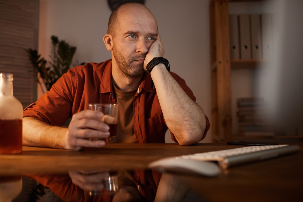 Emotional numbing? Man sitting at the table in front of computer and thinking about something while drinking alcohol