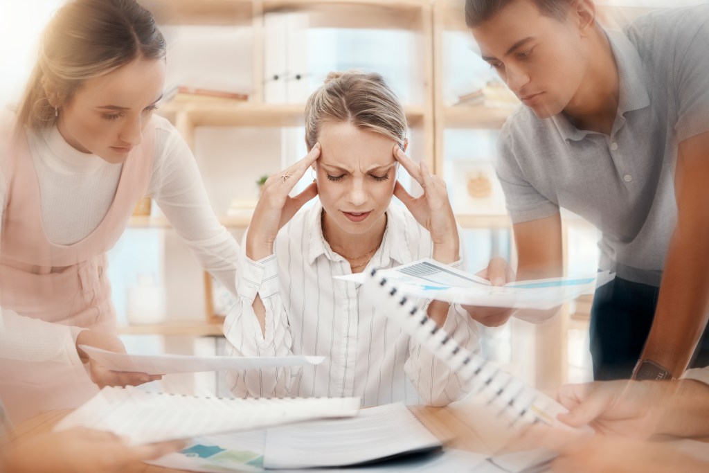 Hidden stressors hiding in plain sight. Woman overwhelmed at work, being bombarded with requests from her fellow workers.