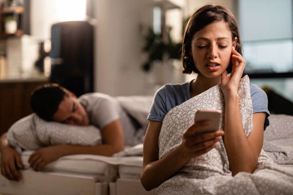 Woman chatting with her AI boyfriend while husband is asleep behind her.