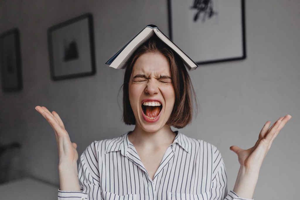 Woman silently screaming with book on her head, frustrated by foreign language anxiety
