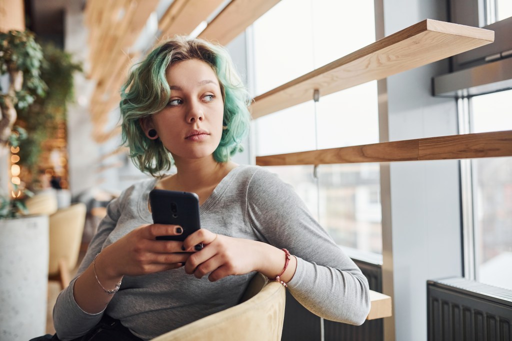 A young woman with wavy, shoulder length blue-green hair holds a cell phone. She is wearing a light gray long sleeve shirt. She is indoors near a large window with a natural wood frame and a radiator. The woman is seated on a tan colored chair. She's talking with her AI mental health app about her social anxiety.