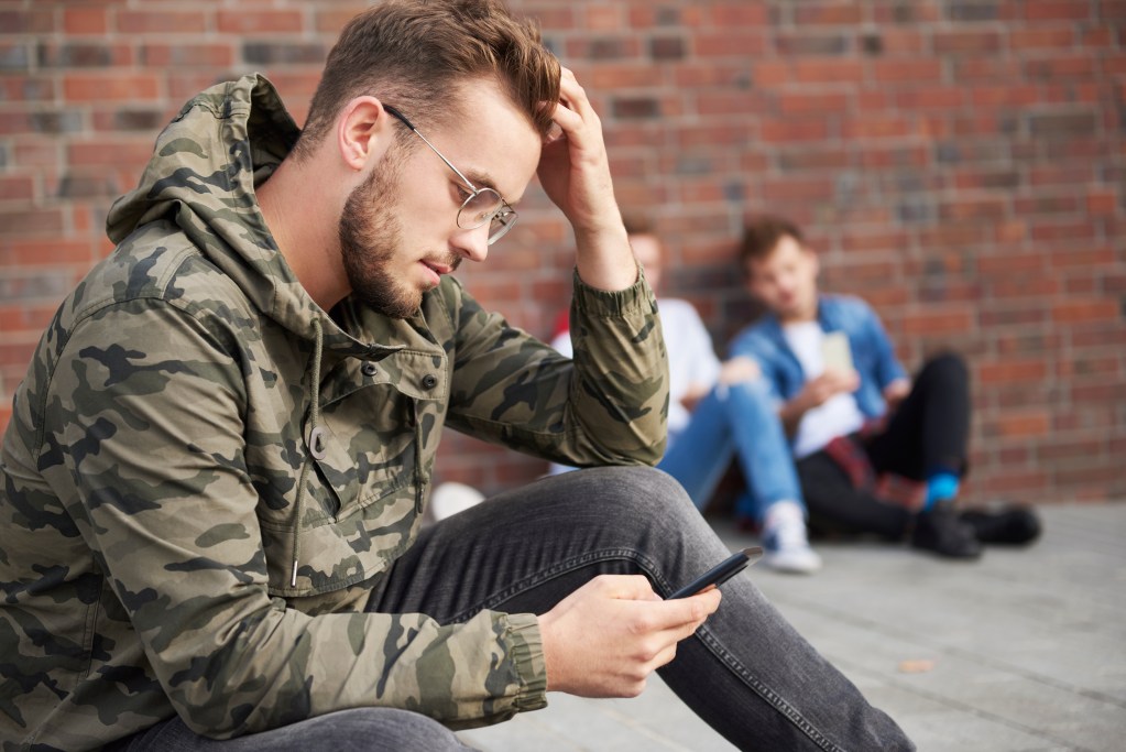 A group of men are sitting in a circle in an interior setting. One man with dark hair, wearing glasses and a striped sweater, sits with his face in his hand, apparently upset. Another man wearing a red beanie and a tan hooded sweatshirt has his hand on his shoulder in a gesture of support. 