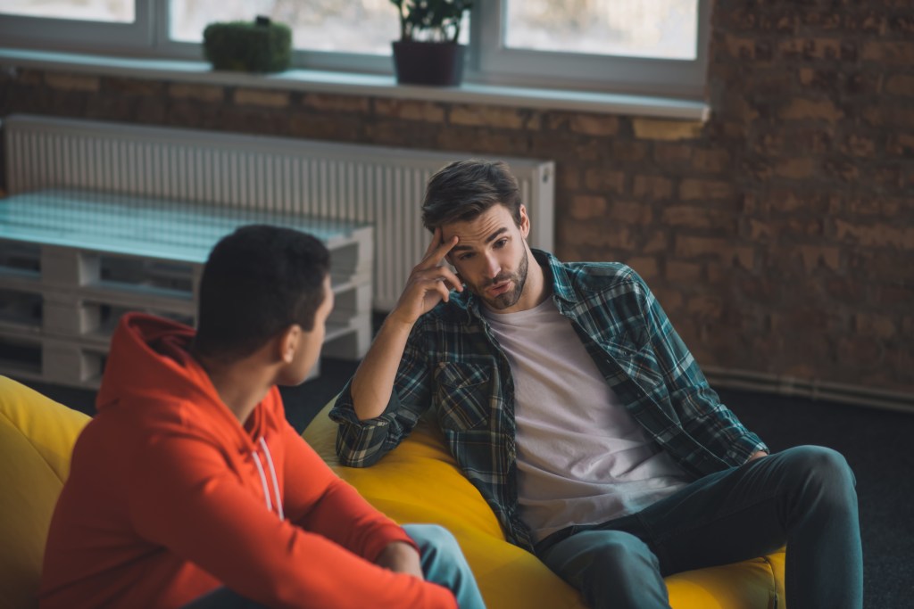 Two men are sitting on a yellow sofa in an urban setting, having a discussion. One man, with dark hair and a beard, is resting his head on his hand. He is wearing a white t-shirt, a green and black flannel shirt, and dark jeans. He appears to be troubled. The other man is turned toward him, listening, and wearing an orange hooded sweatshirt. The warm daylight illuminates a brick wall and modern, minimalist decor. 