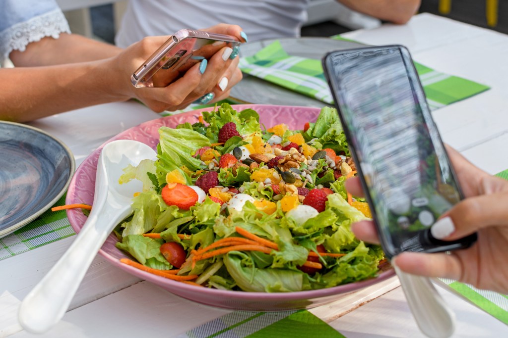 Two people using food apps to photograph a bowl of colorful salad on a white wooden table. One person is holding a rose gold colored smart phone and taking a photo of the salad. The other person is also holding a black smart phone, and appears to be in the process of taking a photo as well. The salad is in a pink bowl and includes lettuce, berries, carrots, fruit, nuts, and other vegetables.