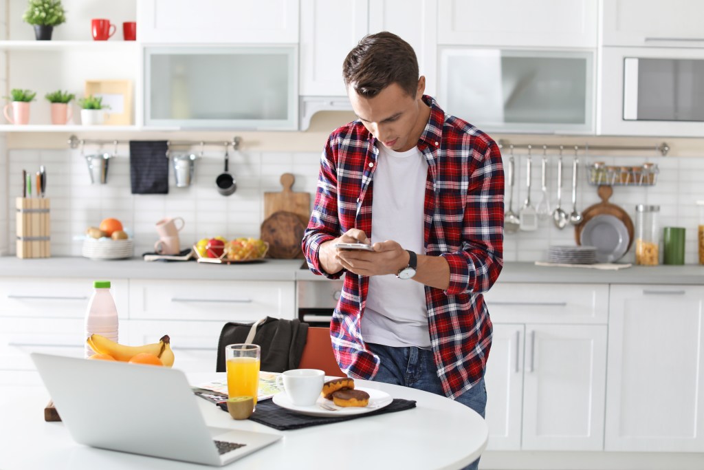 A young adult man with short dark hair is standing at a white table in a bright, white kitchen. He is wearing a white t-shirt and a red and black plaid shirt, and he is looking down at a food app on his smartphone in his hands. On the table is a laptop with more data, a glass of orange juice, a plate with doughnuts, and a cup.