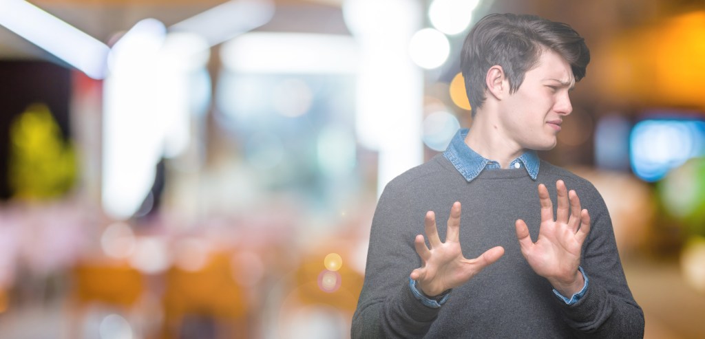 Conversation hangover: A young man with dark hair is posed in a waist up shot making a face of disgust with his hands up. He has a blue denim shirt collar peeking out from under a grey sweater. The background is blurry and shows a pattern of white and orange blobs.