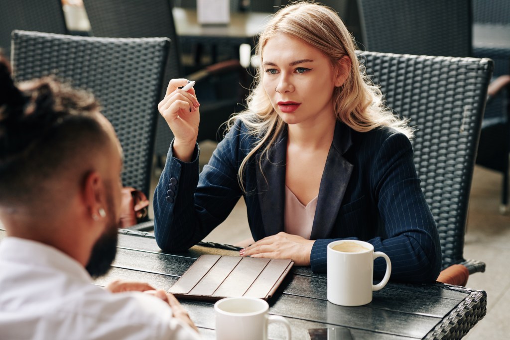 Overcoming Conversational Hangover: A focused woman with wavy blonde hair and red lipstick wearing a navy suit jacket gestures with a pen during a daytime meeting with an associate at a cafe. A mug of coffee, a notepad, and chairs surround the pair. The woman’s expression is self-secure, clear about her boundaries.