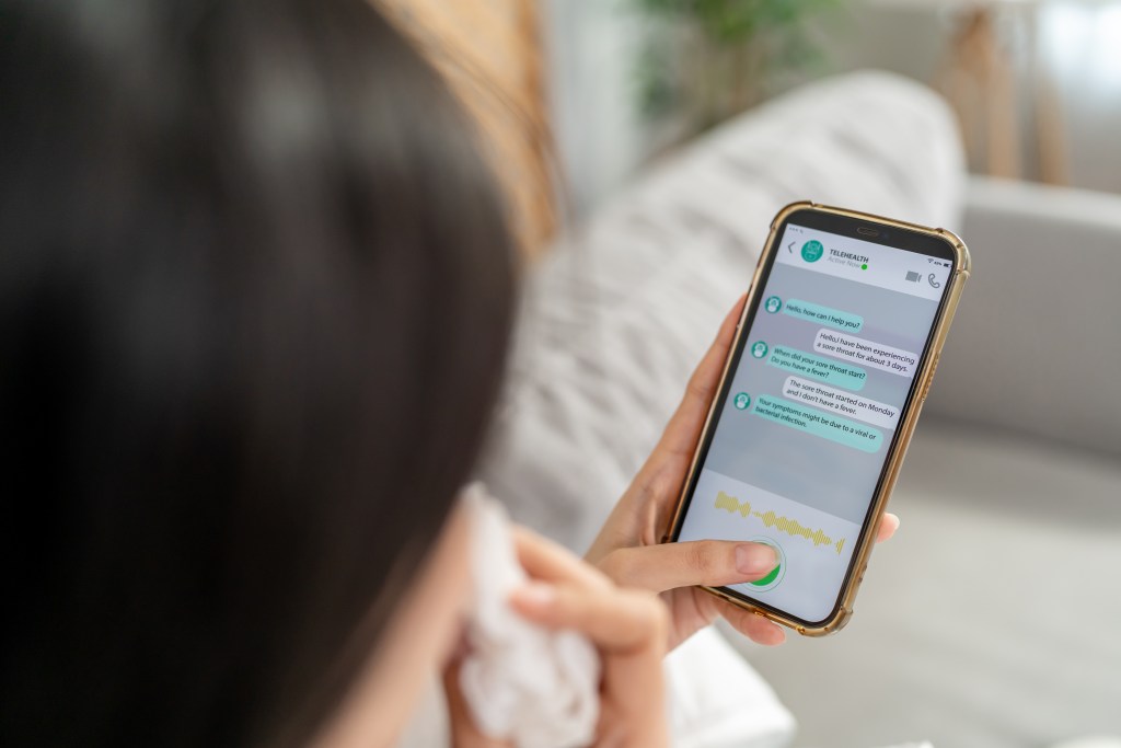 A woman holds a gold-rimmed smartphone with a digital mental health messaging app displayed. The app shows a conversation about a sore throat and possible viral or bacterial infection. The woman is wearing a white garment and appears to be dabbing at her eye with a tissue in her other hand. She is sitting on a couch at home in a well-lit room. 