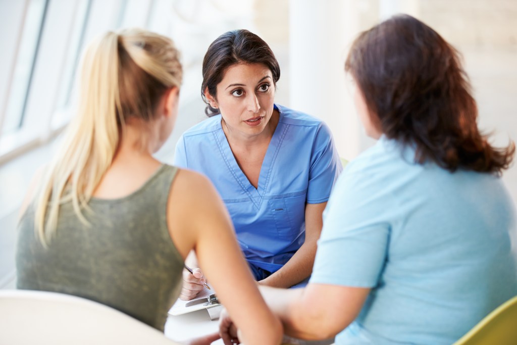 In a bright, clinical setting, an adult mental health nurse in blue scrubs is consulting with two patients. She is looking intently at one of them, and appears to be listening to and answering questions. One patient, with blonde hair pulled back, is holding a pen and clipboard. The other patient has brown hair and is wearing a light blue shirt.