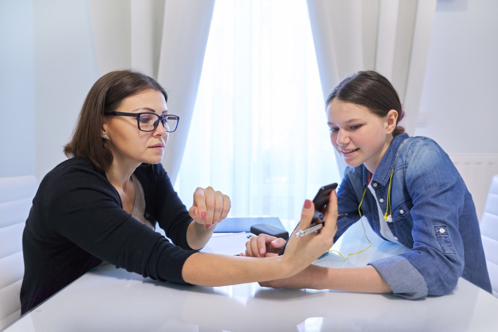 A woman with short brown hair and glasses, wearing a black sweater, sits at a table with a teen who has dark hair. The teen is wearing a denim jacket and has earphones around their neck. They are both looking at a mobile phone with a mental health app that the woman is showing. The setting appears to be indoors, near a window. The mood is light-hearted and casual. 